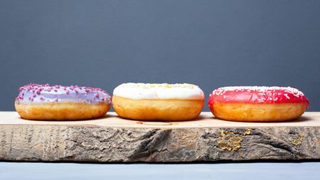 three assorted glazed donuts of different colors on a wooden plank on a gray background.