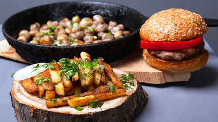 hearty delicious rustic lunch. french fries burger and fried mushrooms champignon in cast iron on forest wooden coasters on a gray background close-up.の写真素材