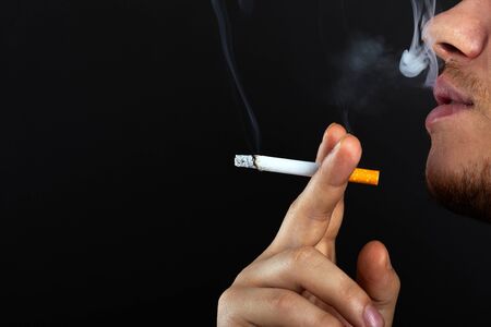A young man holds a cigarette in his hands and exhales smoke on a dark background. harm from smoking...