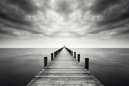 A solitary figure stands at the end of a wooden pier, gazing into the overcast horizon, in a dramatic monochrome scene.の素材