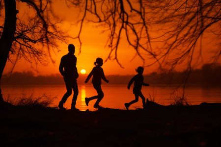 Silhouettes of a family enjoying a walk near the riverside at sunset, with the golden sun reflecting on the water's surface.の素材
