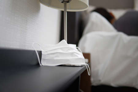 Asian Male sleeping in bed feeling sick with covid-19 symptoms. Non-surgical face mask on nightstand is center of focus. Self quarantine isolationの写真素材