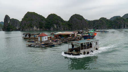 Cat Ba, Vietnam - November 19, 2019 : Fishing boat with clam shellfish passes floating village, near Ha Long Bay, mountain karst, overcast sky.のeditorial素材