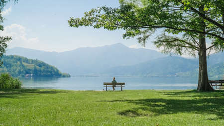 Woman sitting alone on a bench. Attractive girl sits on a wooden bench with amazing view over lake and mountains.Female enjoing a picturesque place. Recreation,peace,mindfulness and relaxation conceptの写真素材