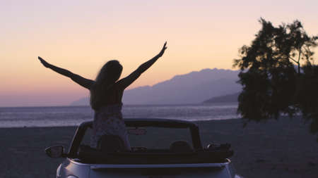 Happy young woman enjoying a ride in a convertible car Back view of woman sitting in cabriolet putting hand on door looking away on background of sea. Travel, freedom, escape and destination concept.の写真素材