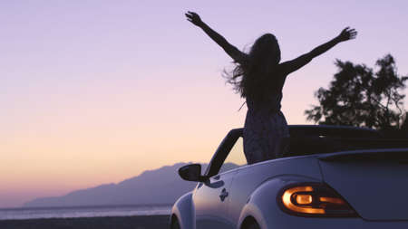 Young pretty womanenjoy white convertible vintage car on exotic island by seaside at sunset....
