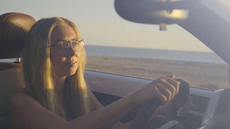 Smiling young blonde woman driving in convertible car along the coastal road in Greece. Pretty...