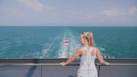 Happy blonde pretty woman standing on the ferry deck and enjoying ocean view on a warm sunny day. Thailand flag waving in the wind. Spectacular Seascape, adventure, destination, exloration concept.の写真素材
