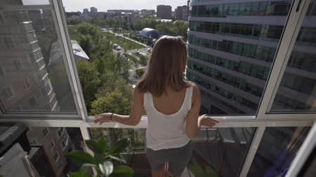 Rear view of a pretty young blonde woman standing in the doorway on the balcony and looking outside into the street on a warm spring day. Covid-19 pandemia, stay home and global lockdown concept.の写真素材