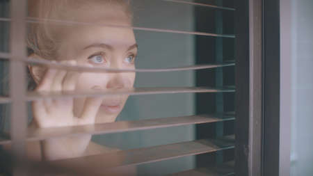 Beautiful young girl separating slats of blinds and looking through windowの写真素材