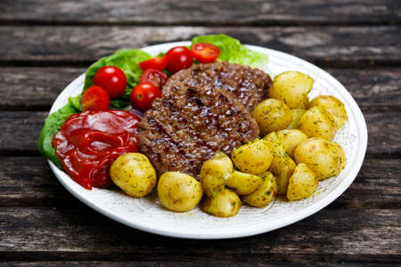 Fresh juicy grill burgers, served with ketchup, young potatoes, tomatoes and vegetables. On wooden table.の写真素材