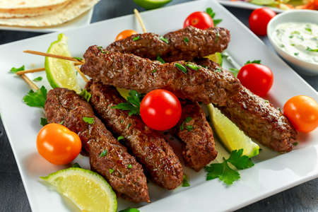 Homemade Kofta kebabs on skewers with pita, lime, vegetables, sweet chili and yogurt sauce on white plate.の写真素材
