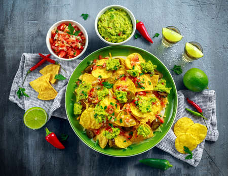 Nachos with cheese, jalapeno peppers, red onion, parsley, tomato, salsa, guacamole sauce and tequila on green plate.の写真素材