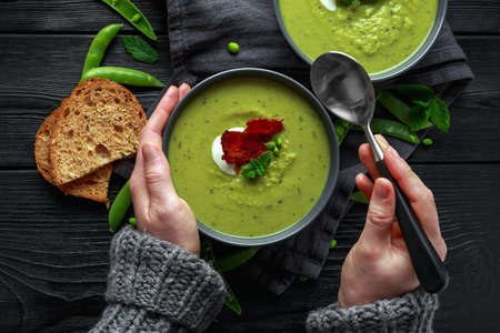 Hands in sweater, pullover holding cosy warming in cold weather. Sweet pea, leek and mint hot soup topped with crunchy bacon and dolop of yogurtの写真素材