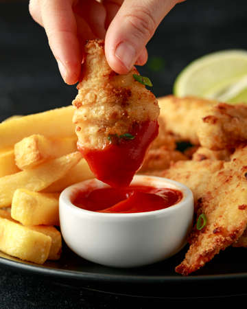 Womans hand dipping Buttermilk chicken nuggets in tomato ketchup served with potato chips and lime...