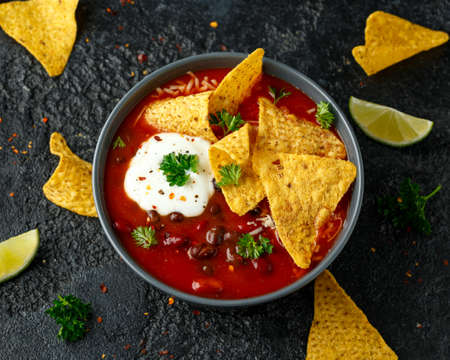 Mexican Chilli bean soup with yogurt, cheese and tortilla chips in black bowl.