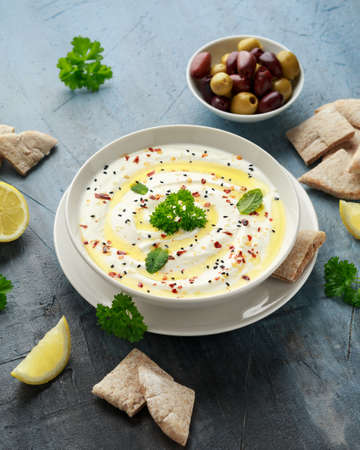 Labneh cream cheese dip with olive oil, salt, herbs served with olives, pita bread in white bowl