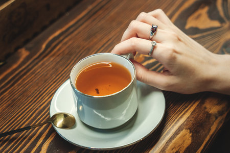 A woman's hand holds a white cup of tea on a wooden table in a cafeの写真素材