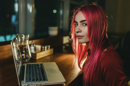 A girl with red hair and a red dress is sitting at a laptop in a cafeの写真素材