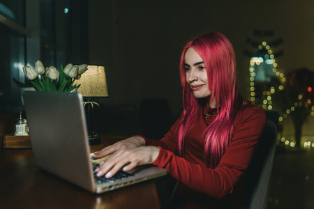 A girl with red hair and a red dress is sitting at a laptop in a cafeの写真素材