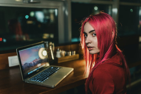 A girl with red hair and a red dress is sitting at a laptop in a cafeの写真素材