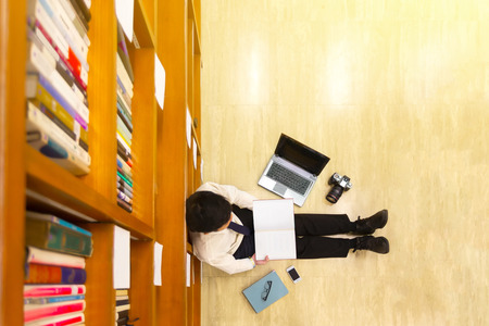 Top view of young business man reading a book in library against multi colored bookshelf in library, keeping a book in his hands and learinng with laptop. Education, Knowledge, Lecture concept.のeditorial素材