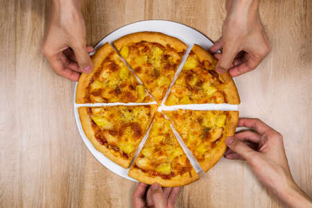 Top view of hands taking slices of delicious and crispy hawaiian pizza topping with mozzarella cheese, pineapple and tomato source. Group of hungry friends sitting at desk and sharing delicious lunch on wooden texture table background.の写真素材