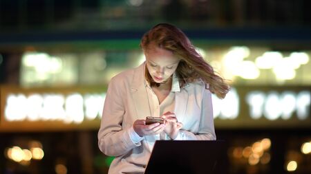 Young beautiful female using smartphone sitting with laptop outs