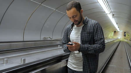 Young happy man surfing internet on his phone while standing on escalator.