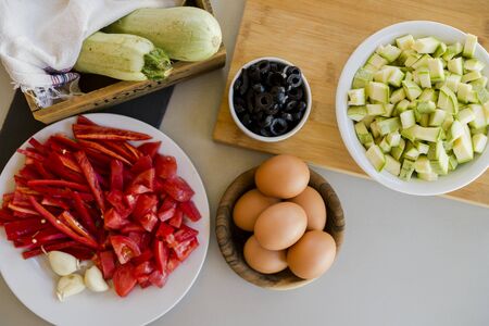 Plates with sliced vegetables and eggs with olives in bowls on kitchen table, cooking process conceptの写真素材