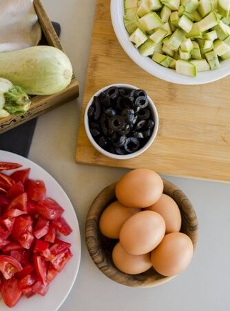 Plates with sliced vegetables and eggs with olives in bowls on kitchen table, cooking process conceptの写真素材