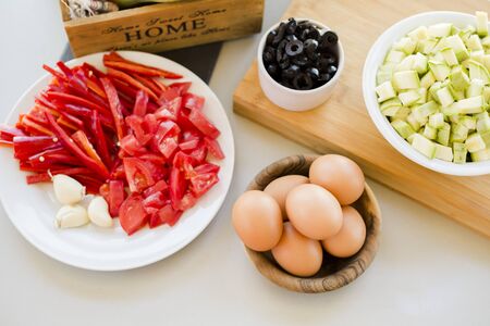 Plates with sliced vegetables and eggs with olives in bowls on kitchen table, cooking process conceptの写真素材