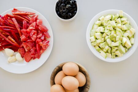 Plates with sliced vegetables and eggs with olives in bowls on kitchen table, cooking process conceptの写真素材
