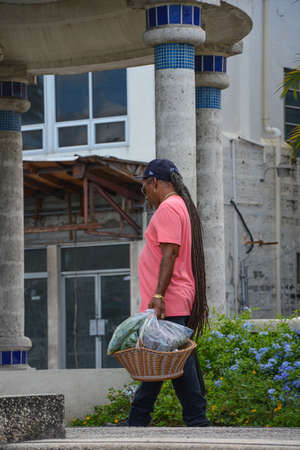 Bridgetown, Barbados, Caribbean - 22 Sept 2018: A man with long hair in a pink T-shirt carries in his hand a wicker basket. Verticalのeditorial素材