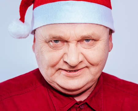Close up portrait of evil aged man in Santa Claus hat and red shirt over white backgroundの写真素材