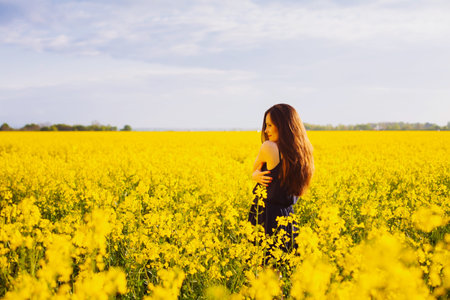 Rear view of young woman with long hair hugging herself on yellow blooming rapeseed fieldの写真素材