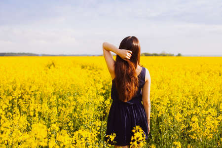 Rear view of young woman with hand in long hair on yellow blooming rapeseed fieldの写真素材