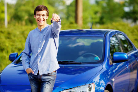 Happy young man in glasses standing in front of his new car and showing car keyの写真素材