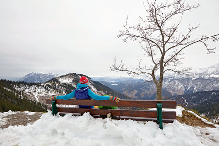 Rear view of female hiker sitting on wooden bench in snowy Alps with view over winter mountain landscapeの写真素材