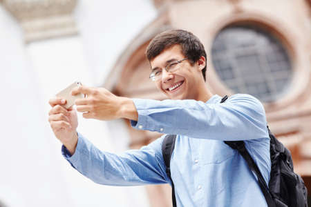Smiling young man in glasses taking photo of himself with smartphone near cathedral during vacationの写真素材