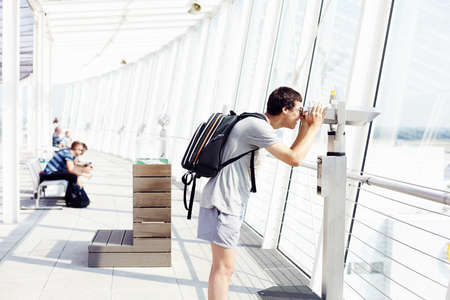 Profile of smiling young man with backpack looking through coin operated telescope near window in airportの写真素材