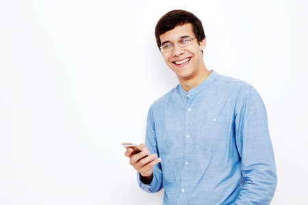 Young hispanic man wearing jeans shirt and glasses holding smartphone in his hand and smiling against white wall - communication conceptの写真素材