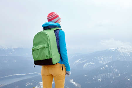 Back view of young woman wearing pink hat, blue jacket, green backpack and yellow pants standing against winter mountains - adventure conceptの写真素材