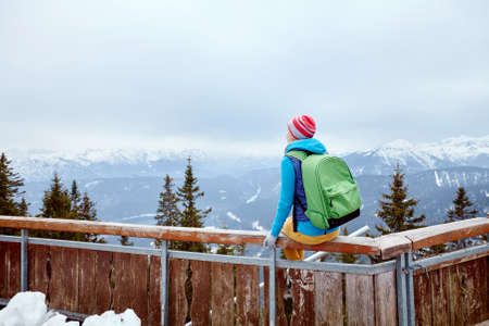Back view of young woman wearing pink hat, blue jacket, green backpack and yellow pants sitting on wooden fence against winter mountains - adventure conceptの写真素材