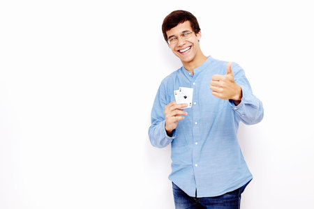 Young hispanic man wearing jeans and glasses holding two aces (hearts and clubs) in his hand and showing thumb up hand gesture with smile against white wall - gambling conceptの写真素材