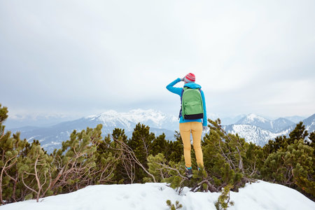 Back view of young woman wearing pink hat, blue jacket, green backpack, yellow pants and hiking boots standing against winter mountain valley and looking far covering her eyes by hand - adventure conceptの写真素材