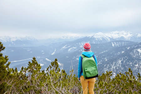 Back view of young woman wearing pink hat, blue jacket, green backpack and yellow pants standing against winter mountain valley - adventure conceptの写真素材