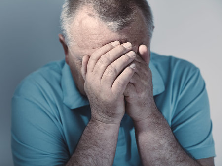 Dramatic cool toned close up portrait of aged man sitting with hands on his face against white wall...