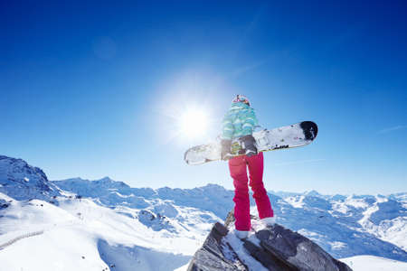 Back view of female snowboarder wearing colorful helmet, blue jacket, grey gloves and pink pants standing with snowboard in one hand and enjoying sunny alpine mountain landscape - winter sports conceptの写真素材