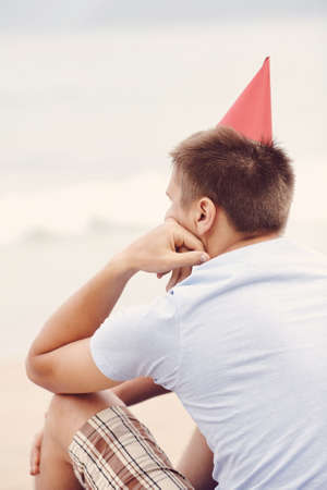 Retro colored back view portrait of young man wearing red party hat, blues t-shirt and checkered shorts celebrating birthday sitting alone with hand under his chin on beach near tropical seaの写真素材